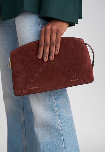 Μπορντό σουέντ τσάντα χιαστί με διαγώνια ραφή, χρυσό φερμουάρ και μικρό ιμάντα καρπού, κρατημένη από ένα χέρι εναντίον ανοιχτόχρωμου τζην.