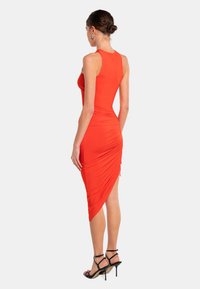 Rotes, figurbetontes Kleid mit runder Rückenausschnitt, asymmetrischem Saum und gerafften Details. Getragen mit schwarzen, samtigen, hochhackigen Sandalen.