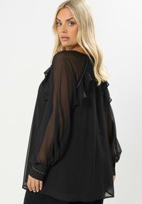 Blusa nera in chiffon con dettagli arricciati e maniche lunghe a sbuffo. Accentuata con piccoli borchiette lungo i polsini e sul retro.