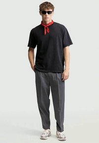 Mand iført sort t-shirt, grå plisserede bukser, hvide sneakers, rød halsedisse og sorte solbriller, stående med hænderne i lommerne.