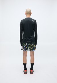 Bald person som står med ryggen til iført en svart langermet skjorte, blomstrete shorts, svarte sokker og oransje-svarte sko på en ensfarget bakgrunn.