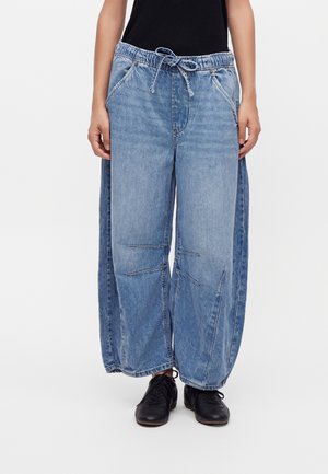 Personne portant un pantalon en denim bleu clair ample avec une taille élastique et un cordon, associé à des chaussures noires et un haut noir.