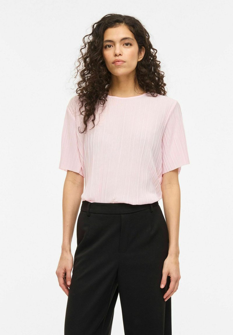 Žena s kučeravými vlasmi v svetloružovej krátkoročnej textúrovanej blúzke a čiernych nohaviciach s vysokým pásom, stojaca pred jednoduchým pozadím.