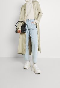 Person som bär en beige trenchcoat, en krämfärgad polotröja, ljusblå jeans, vita chunky sneakers och håller en svart handväska.