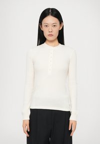Top a maniche lunghe di colore crema realizzato in tessuto a costine, con scollatura rotonda e dettagli con bottoni lungo la parte anteriore. Design aderente.