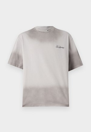 Fiatal férfi, zsebében tartott kézzel, laza szürke pólót, fekete pliszírozott térd alatti szoknyát, fekete zoknit és fekete tornacipőt visel.