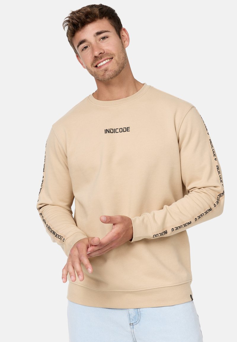 Sudadera beige de manga larga, con escote redondeado, logo 'INDICODE' en el pecho y las mangas, tejido suave y puños de canalé.