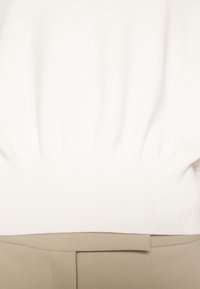 Bílý pletený top s žebrovaným lemem, v kombinaci s béžovými látkovými kalhotami s hladkou texturou a jednoduchým zapínáním v pase.