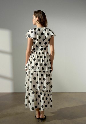 Frau trägt ein weißes mehrlagiges Kleid mit großen schwarzen Punkten, steht drinnen vor einer schlichten grauen Wand und blickt von der Kamera weg.