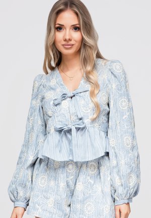Jeune femme aux longs cheveux blonds portant un chemisier bleu brodé de fleurs et un short assorti avec des détails de nœud à l'avant.