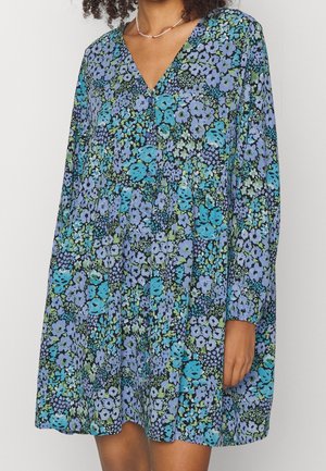 Kobieta w sukience z długim rękawem i dekoltem w serek, z niebiesko-fioletowo-zielonym kwiatowym wzorem i guzikami z przodu.