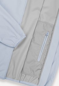 Világosszürke kabát belső része cipzáras belső zsebbel és elasztikus mandzsetta részlettel az ujjon, részben kinyitva bemutatva.