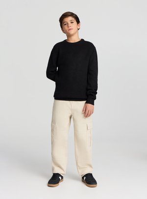 Maglione nero a costine con maniche lunghe, abbinato a pantaloni cargo beige con tasche laterali. Scarpe da ginnastica nere con suole marroni.