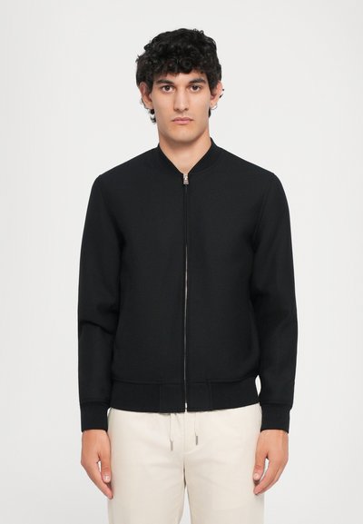 Giacca bomber nera con zip frontale, polsini e orlo a costine. Texture liscia e design minimalista, abbinata a pantaloni chiari.