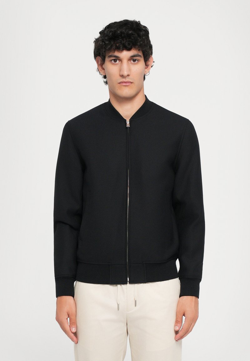 Melna zip-up bomber jaka ar ribotu apkakli un piedurknēm. Gluda tekstūra, pieguļošs izskats un minimālistisks dizains. Uzvilkta virs gaišas krāsas biksēm.