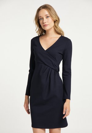 Jeune femme aux cheveux blonds ondulés portant une robe portefeuille à manches longues, longueur genou, bleu marine foncé sur un fond clair.