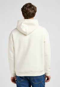 Hoodie color crema con cappuccio regolabile, texture morbida e polsini a coste. Abbinato a jeans blu, presenta una vestibilità comoda e uno stile informale.