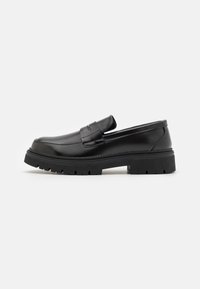 Black leather loafers with a flat, ridged sole. Features a stitched design and a slip-on style with a decorative strap across the front.