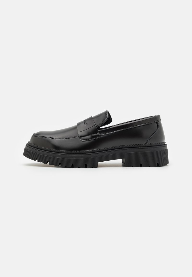 Black leather loafers with a flat, ridged sole. Features a stitched design and a slip-on style with a decorative strap across the front.