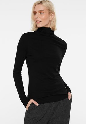 Blonde woman wearing a black turtleneck and gray pants with hands in pockets, looking to the side against a plain background.