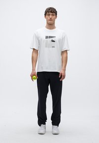 Λευκό βαμβακερό T-shirt με εκτυπωμένο σχέδιο, συνδυασμένο με μαύρο παντελόνι και γκρι αθλητικά παπούτσια. Το μοντέλο κρατά μια κίτρινη μπάλα του τένις.