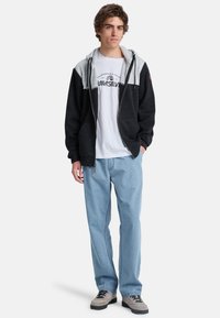 Felpa con cappuccio caratterizzata da blocchi di colore grigi e neri, con zip frontale, tasche laterali e logo su una maglietta bianca. Jeans azzurri.