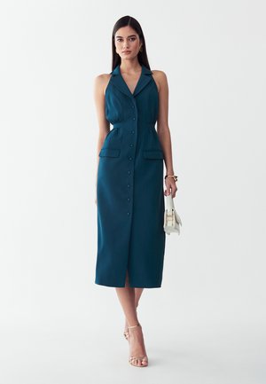 Vrouw draagt een mouwloze teal midi-jurk met knopen en kraag, houdt een witte handtas vast en draagt gouden sandalen en creool oorbellen.