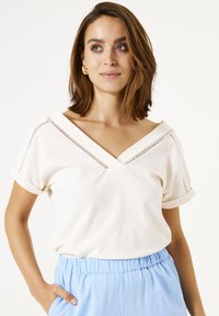 Haut blanc à manches courtes avec un col en V, doté d'un tissu texturé et de coutures décoratives le long de l'encolure. Accompagné d'un pantalon bleu clair.