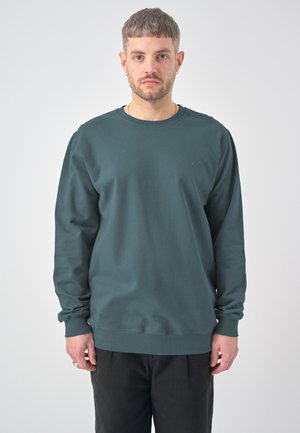 Mann mit kurzen Haaren und Bart, der einen dunkelgrünen langärmeligen Pullover und schwarze Hose trägt und vor einem einfarbigen hellen Hintergrund steht.