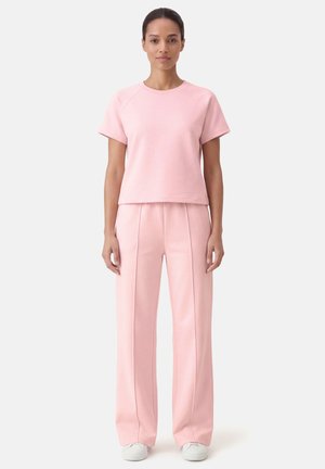 Nainen, jolla on vaaleanpunainen lyhythihainen yläosa ja siihen sopivat leveälahkeiset housut, seisoo suorana yksivärisen vaalean taustan edessä.