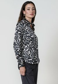Donna con capelli lunghi posa indossando una blusa leopardata e pantaloni grigi, mostrando un'espressione sicura di sé su uno sfondo neutro.