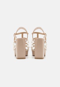 Sandalias de tacón alto beige con tacones de bloque grueso, múltiples correas y tachuelas decorativas; hechas de un material sintético texturizado.