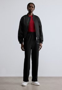 Czarna kurtka typu bomber, czerwony obcisły top, czarne spodnie dresowe z logo, białe trampki. Gładkie tekstury i kontrastujące kolory na całej powierzchni.