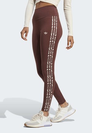 Femme portant des leggings marron taille haute avec des bandes latérales blanches et à rayures zèbres, ainsi que des baskets beiges Adidas.