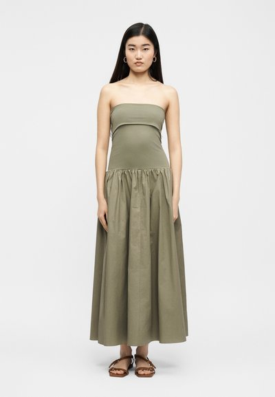 Γυναίκα με μακριά σκουρόχρωμα μαλλιά φοράει ελιάς μακριά φόρεμα χωρίς τιράντες και καφέ επίπεδα σανδάλια, όρθια μπροστά σε απλό ανοιχτό φόντο.