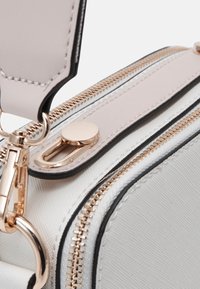 Kompaktní bílá taška s текстurovaným povrchem, vybavená zlatou kováním, dvěma zipy a odnímatelným popruhem. Ozdobena černými lemy.