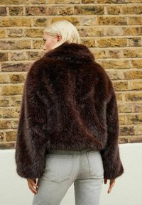 Veste en fausse fourrure brune avec un col montant, présentant une surface texturée et duveteuse, et une coupe cropped, assortie à un jean gris clair.