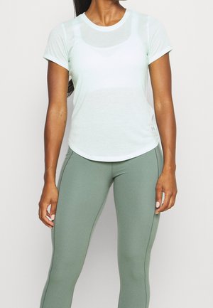 Kobieta w jasno-miętowej sportowej koszulce z krótkim rękawem i dopasowanych legginsach w kolorze szałwii, stojąca na jasnym, jednolitym tle.