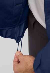 Giacca blu navy con chiusura a coulisse all'orlo, foderatura interna testurizzata e un primo piano di una mano che regola il cordino.