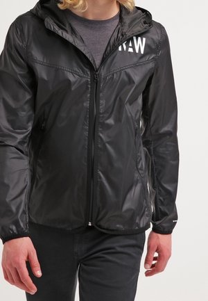 Person trägt eine schwarze Kapuzenjacke mit Reißverschluss und weißem "RAW"-Schriftzug auf der Brust, kombiniert mit schwarzen Hosen.