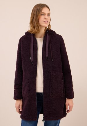 Femme portant une longue veste en polaire violet foncé avec des poches avant, une capuche et des cordons, sur un haut clair et un jean bleu.