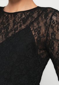 Črna čipkasta majica z rožnim vzorcem, prosojno teksturo, okroglim izrezom in dolgimi rokavi z dodatkom enobarvnega blaga.