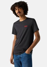 Mand med krøllet hår iført en sort Levi's t-shirt og blå jeans, stående med den ene hånd i lommen foran en ensfarvet baggrund.