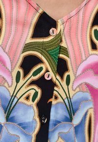 Zbliżenie bluzki z kwiatowym wzorem w pastelowych odcieniach różu, niebieskiego, zielonego i czarnego, z małymi okrągłymi, pokrytymi tkaniną guzikami.