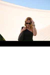 Femme en robe noire asymétrique et lunettes de soleil tenant un sac noir, ajustant ses cheveux devant un fond en tissu clair en extérieur.