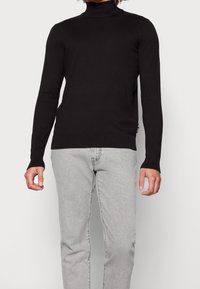 Musta poolokauluspaita, jossa pitkät hihat, resorit hihansuissa ja vartalonmyötäinen leikkaus, puettuna vaaleanharmaiden farkkujen päälle. Sileä kangaspinta.