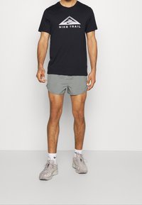 Hombre vestido con una camiseta negra de Nike Trail, pantalones cortos de running grises, calcetines blancos de Nike y zapatillas ligeras grises, de pie contra un fondo blanco.