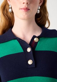 Gestreiftes Polohemd in Marineblau und Grün mit Kragen, drei goldenen Knöpfen und einem roten Etikett; glatte Stricktextur.