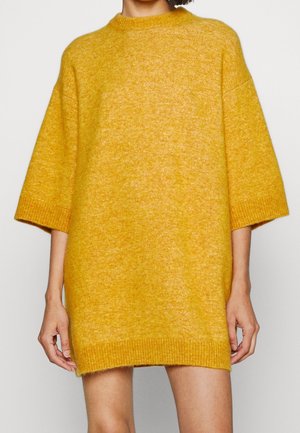 Vrouw draagt een oversized mosterdgele gebreide sweaterjurk met elflengte mouwen en geribbelde omslagen.