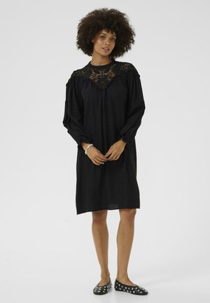 Black dress with long, puffed sleeves and a lace-trimmed yoke detail. Soft, flowing fabric; knee-length design. Worn with black studded flats.
